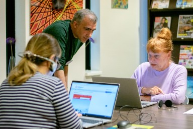 Een oude man met een koptelefoon op zit achter een laptop. Een andere man wijst iets aan op de laptop. 